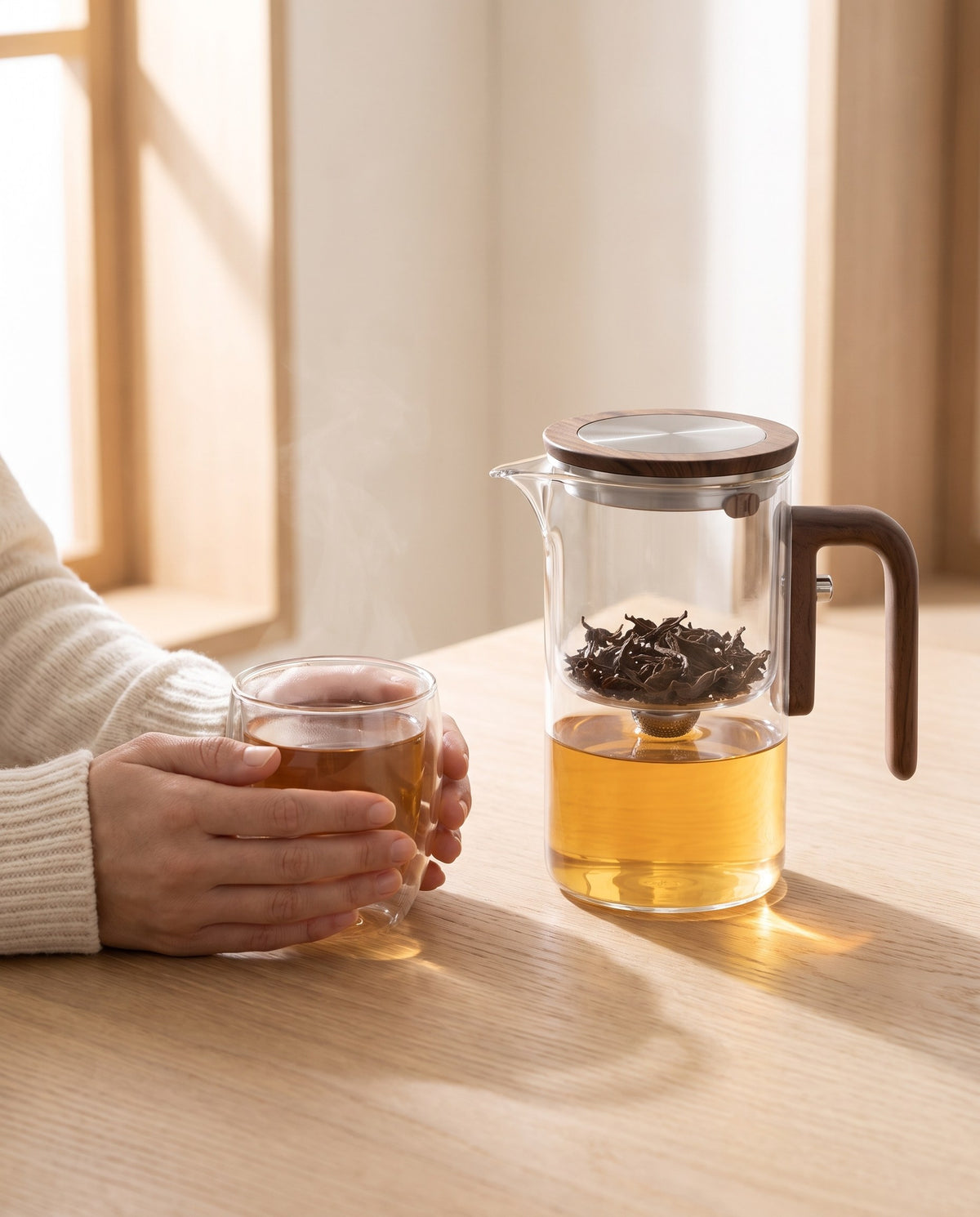 Hände halten eine Glastasse Tee, daneben die Teekanne
